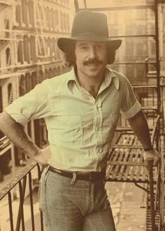 a man standing on a balcony with his hands on his hips and wearing a hat