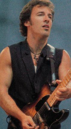 a man playing an electric guitar while wearing a black vest and neck tie, with his eyes closed