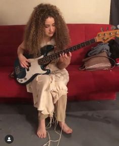 a woman sitting on a red couch while playing a bass guitar with her legs crossed