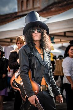 a man with long hair wearing a black hat and leather jacket playing an electric guitar