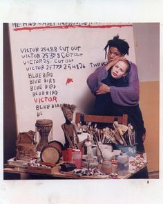a woman and child standing in front of a white board with writing on it, surrounded by art supplies