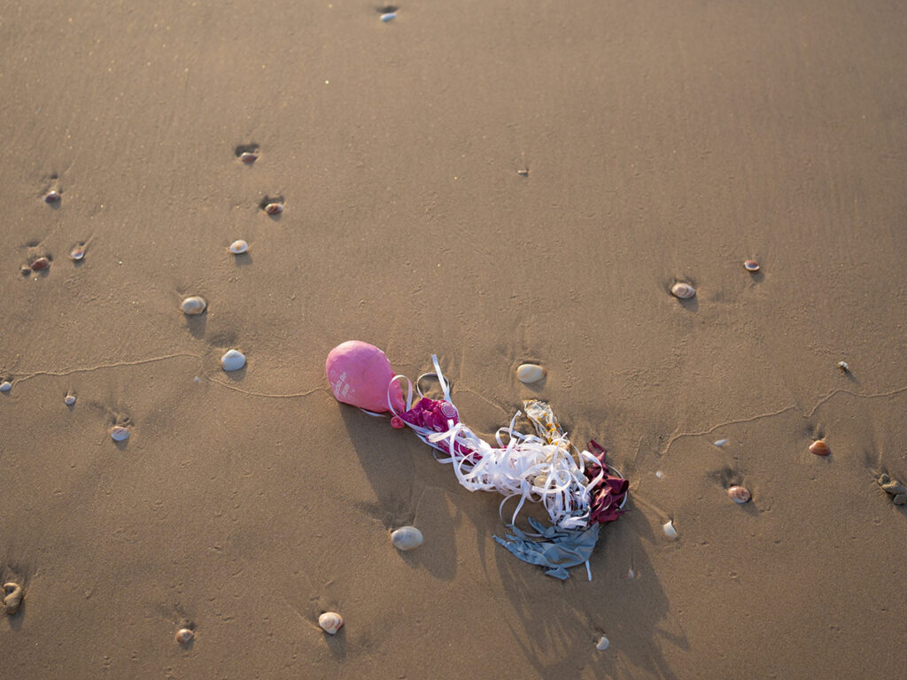 A deflated pink balloon with white ribbon on a sandy beach. White pebbles are scattered around in the sand.