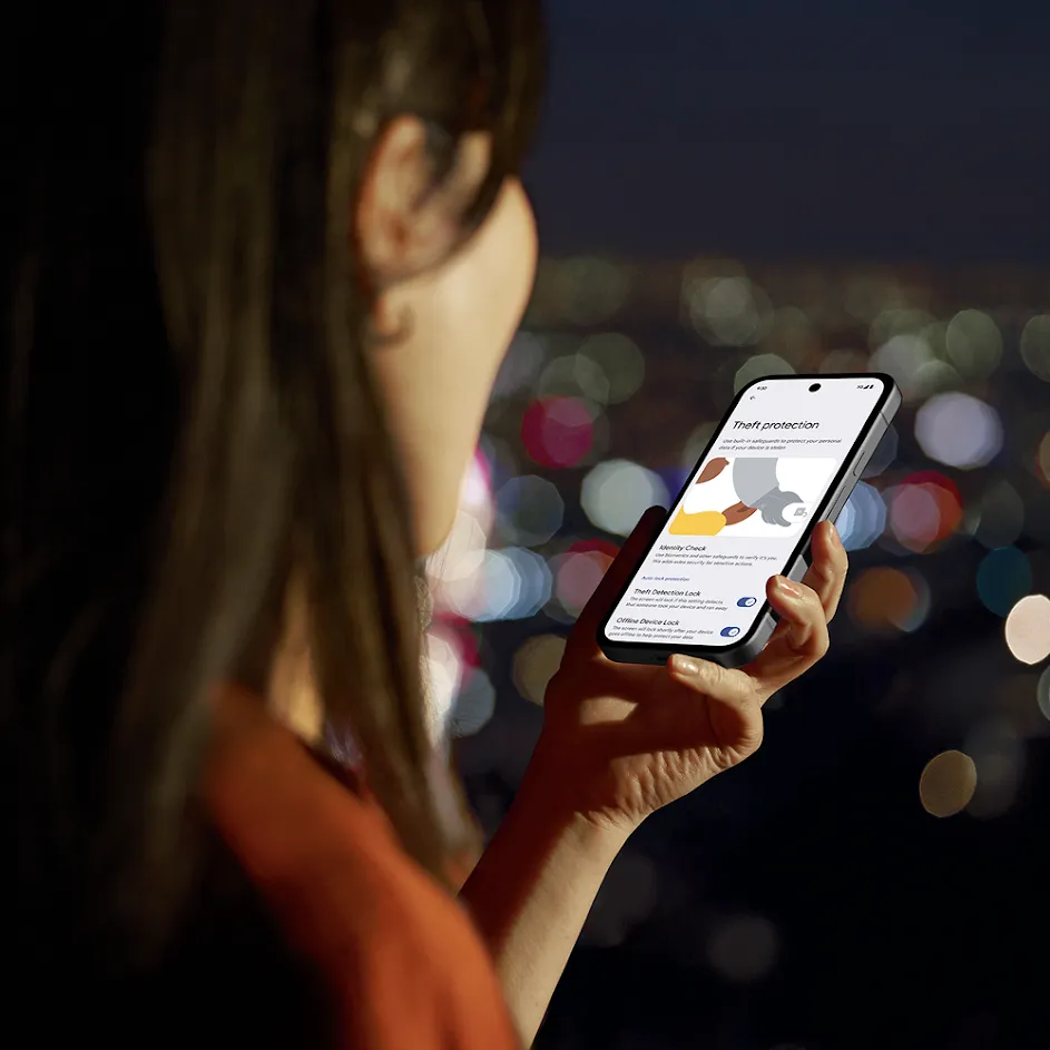 Over-the-shoulder photograph with shallow depth of field, where a woman outside at night with city lights in the background holds up her phone while reading an informational screen about theft protection.
