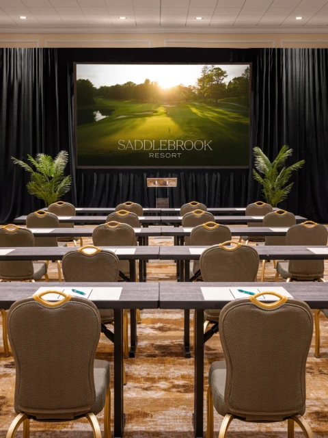 A conference room set up with rows of tables and chairs facing a screen displaying 